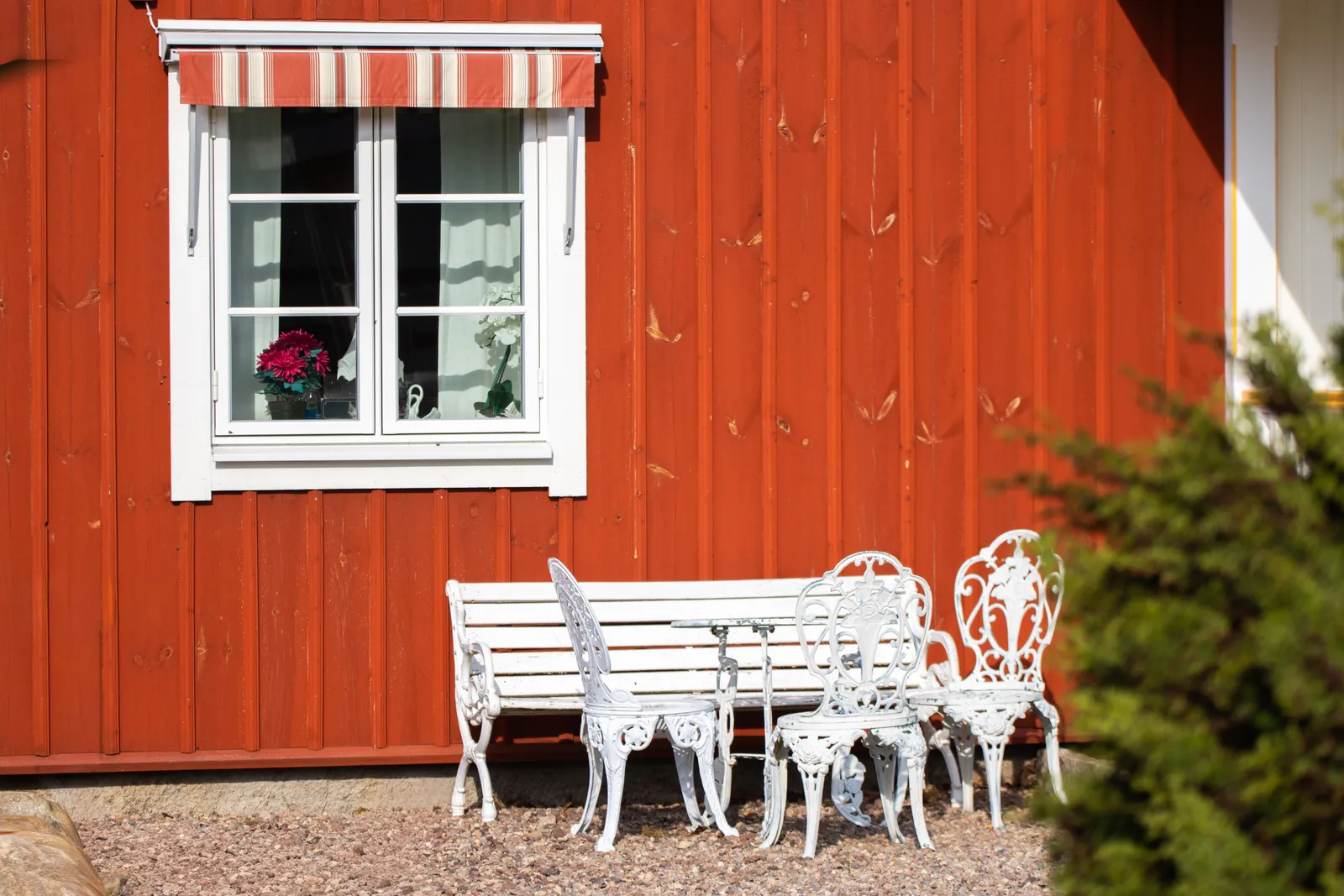Faluröd trävägg med randig markis över fönstret och vita trädgårdsmöbler i solen